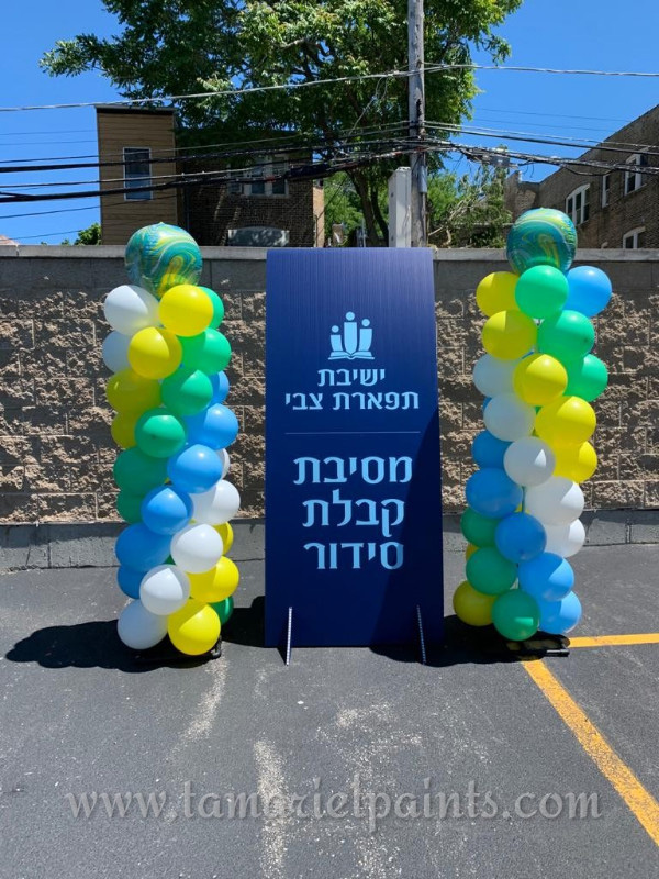 A photo of 2 spiral columns of white, yellow, green and blue ballons on either side of a sign